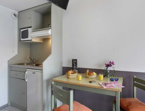 Una kitchenette compacta con microondas, pileta y placard. Una mesa con una cesta de pan, fruta y vasos de jugo está apoyada contra una pared.