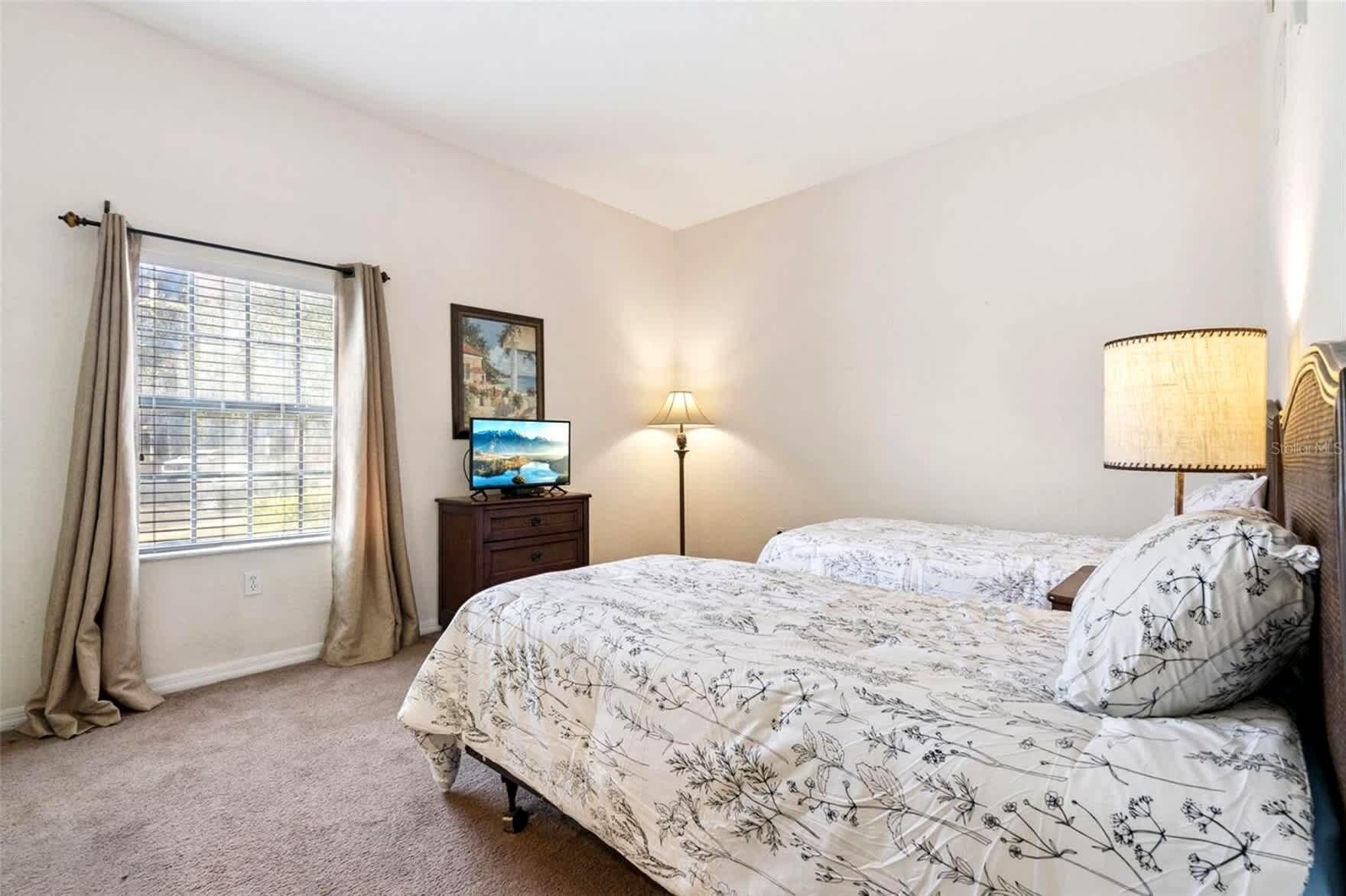 A bedroom with two beds, a TV on a dresser, a lamp, and a window with curtains.