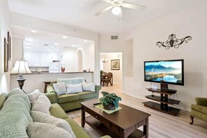 A living room with a sofa, a coffee table with a plant, a TV on a stand, a dining area with a table and chairs, and a kitchen in the background. - Sunlit Escape Resort Stay (Kissimmee)
