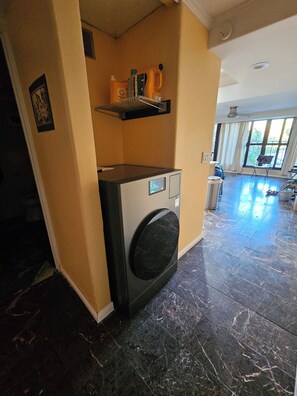 A laundry area with a washing machine, a shelf with cleaning supplies, and a large window providing natural light. - Maui Get-away.  1 BR condo with pool (Lahaina)