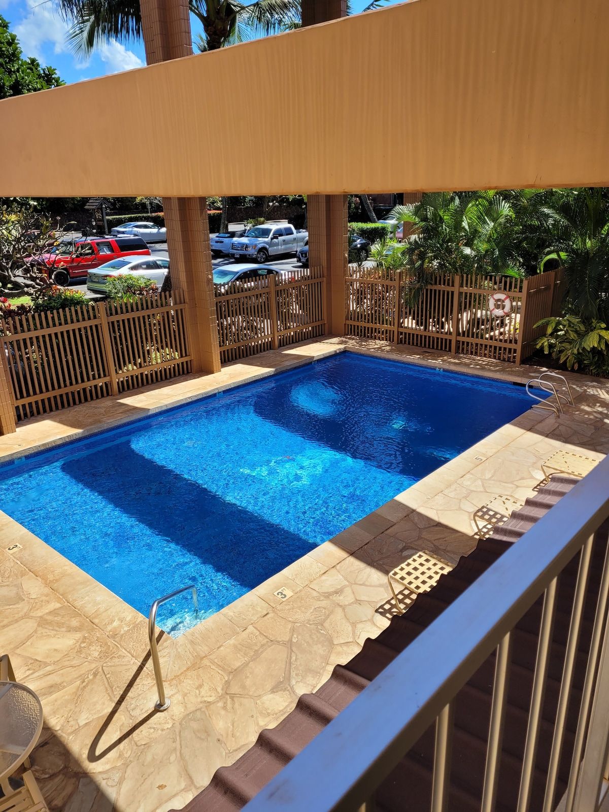Una piscina rectangular con agua cristalina azul, rodeada por una cerca de madera y un patio con baldosas.