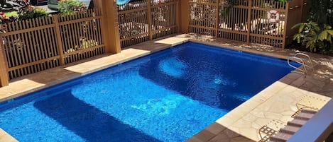 Una piscina rectangular con agua cristalina azul, rodeada por una cerca de madera y un patio con baldosas.