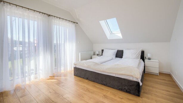 Un dormitorio con una cama grande, un tragaluz, una ventana con cortinas traslúcidas y suelo de madera.