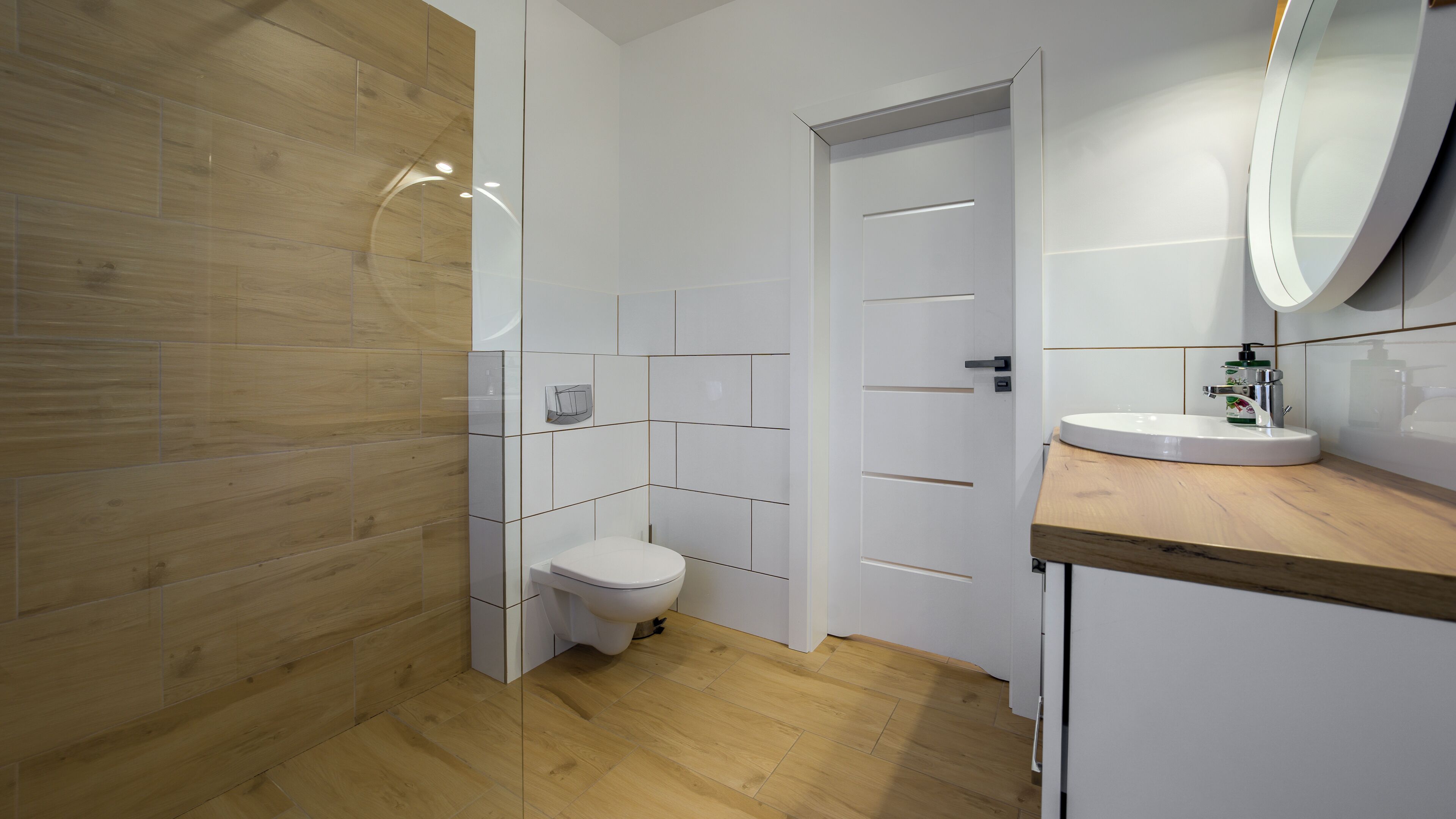 A modern bathroom with a wooden floor, white tiles, a toilet, a sink with a mirror, and a shower area.