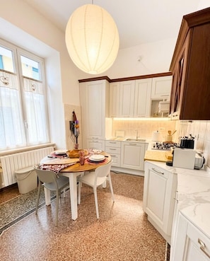 Cozinha com armários brancos, mesa de jantar com cadeiras, luminária pendente e janela com cortinas.