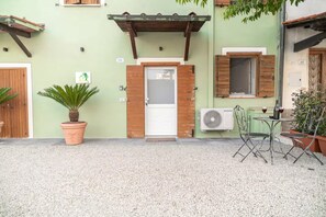 Ein Gebäude mit grüner Fassade, einer Holztür, einem Blumentopf, einem kleinen Tisch mit Stühlen und einem Ventilator.