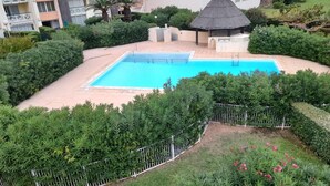 En swimmingpool omgiven av grönska, en lusthus med halmtak och en hög byggnad i bakgrunden.