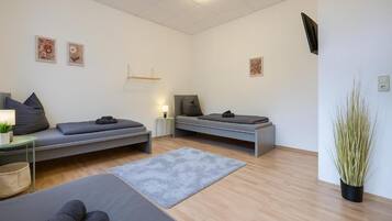 A modern, minimalist room with two beds, a small table with a lamp, a large plant, and artwork on the walls.