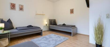 A modern, minimalist room with two beds, a small table with a lamp, a large plant, and artwork on the walls.