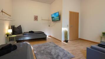 Ein modernes Wohnzimmer mit einer Couch, einem kleinen Tisch mit Lampe, einem an der Wand montierten Fernseher und einer Tür.