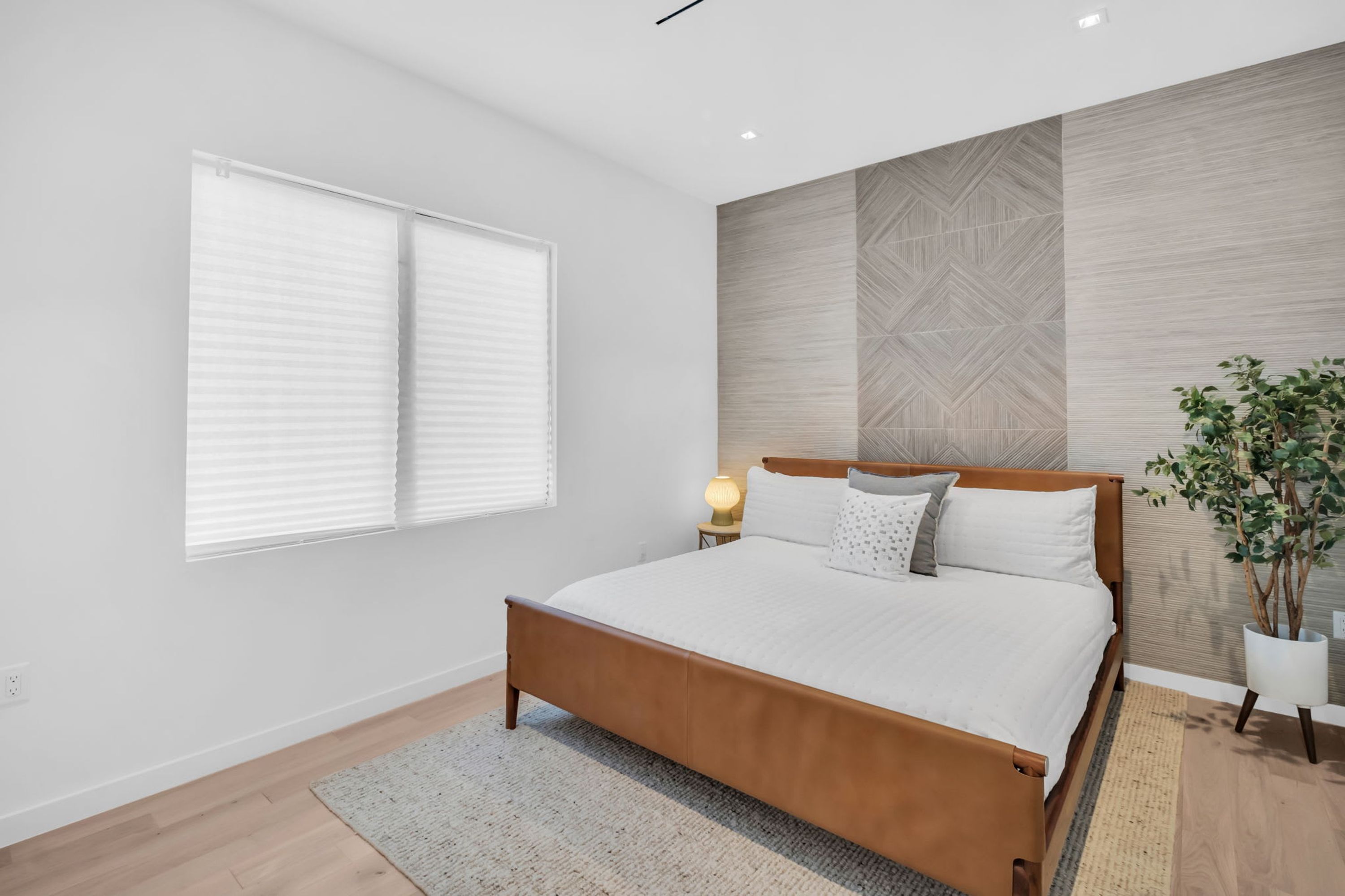 Une chambre à coucher avec un lit en bois, une literie blanche, une table de chevet avec une lampe, une plante en pot et une fenêtre avec des stores.