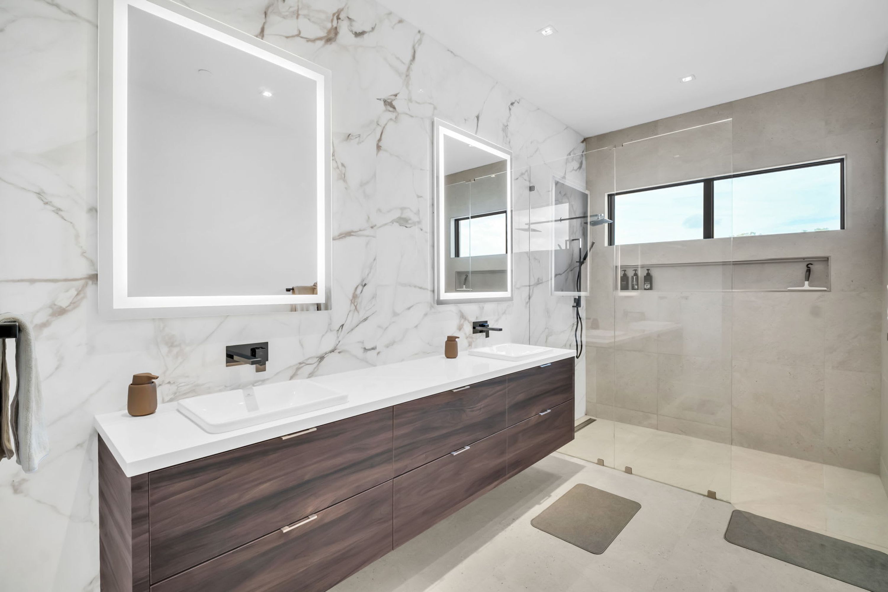 Une salle de bain moderne avec un mur en marbre, un grand miroir, un meuble-lavabo à deux vasques et une cabine de douche en verre.