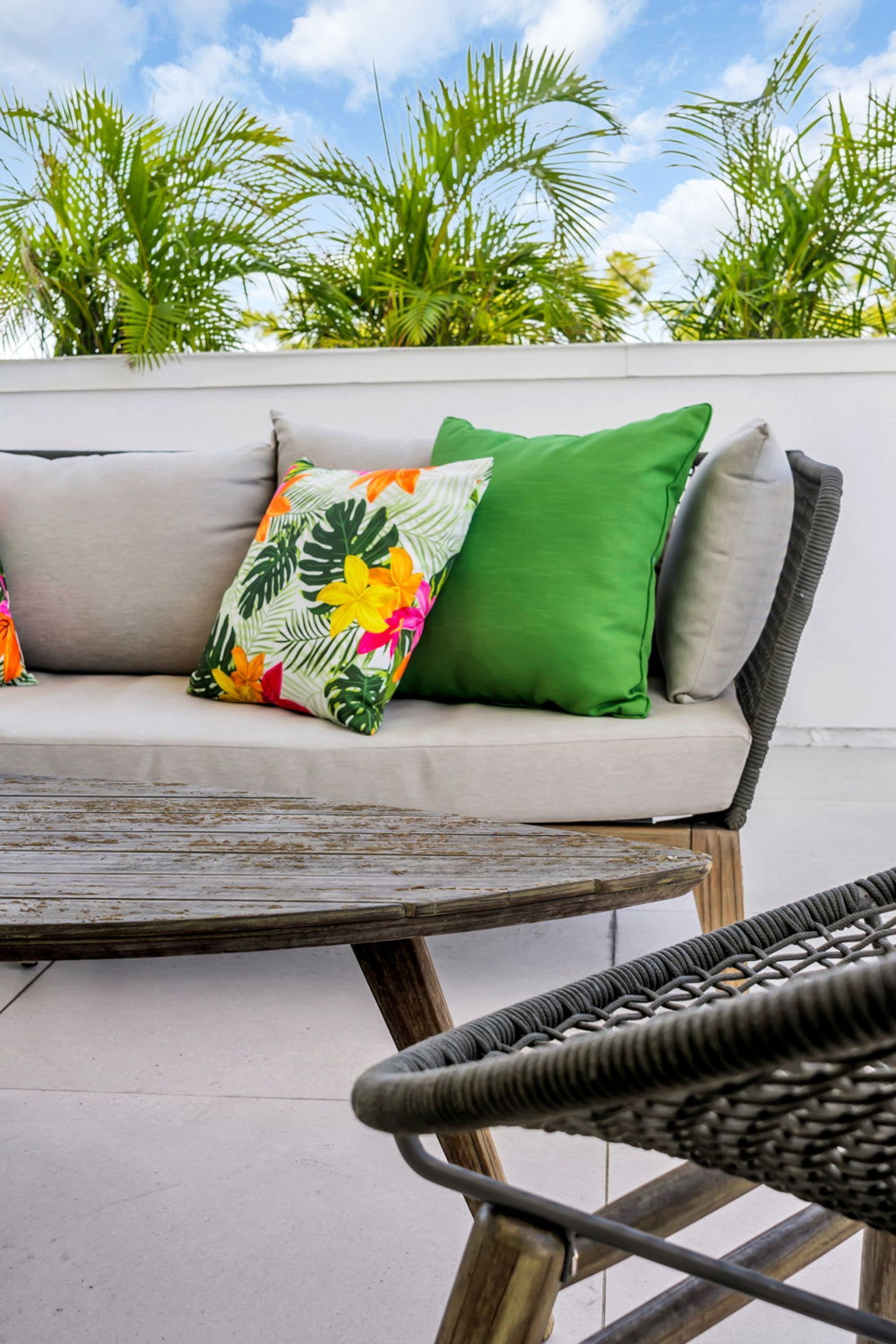 A sofa with tropical patterned pillows, a wicker chair, and a view of palm trees.