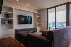 A modern living room with a flat-screen TV, built-in shelves, a glass coffee table, and a large window with a city view. - w Upscale 2BR Duplex w Balcony in Miraflores (Lima)