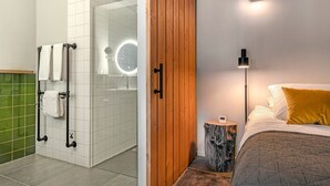 Un baño con cabina de ducha de vidrio, puerta de madera, mesita de noche con un reloj y cama con almohada amarilla.