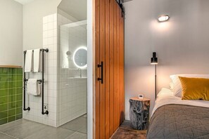 A bathroom with a glass shower enclosure, a wooden door, a bedside table with a clock, and a bed with a yellow pillow. - Lennepserf - De Achterhoek (Veessen)