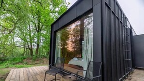 A modern cabin with a wooden deck, two black metal chairs, a woven basket, and a large window overlooking a forest.