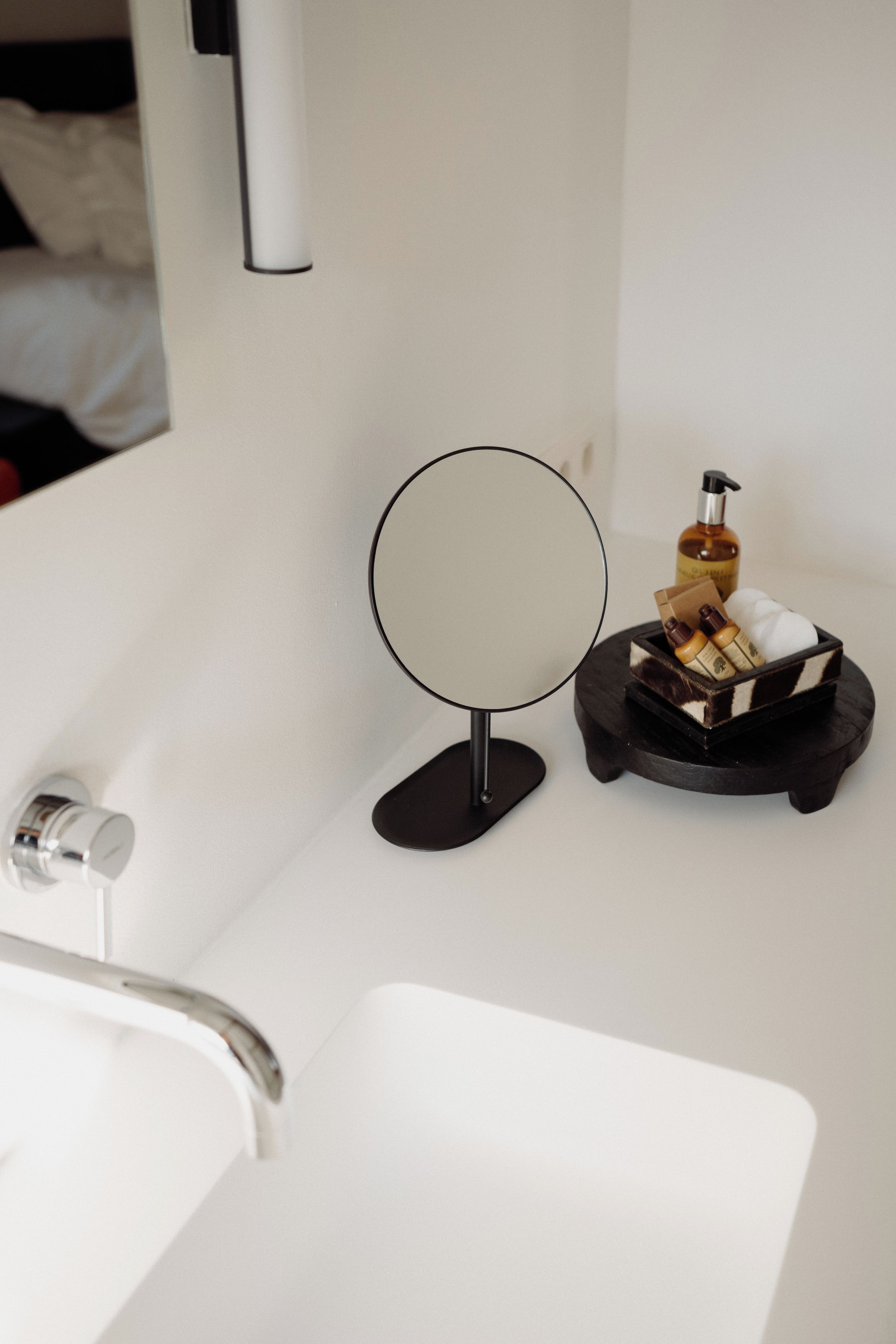 Un bagno con un lavandino bianco, uno specchio rotondo e un piccolo vassoio di legno con articoli da toilette.