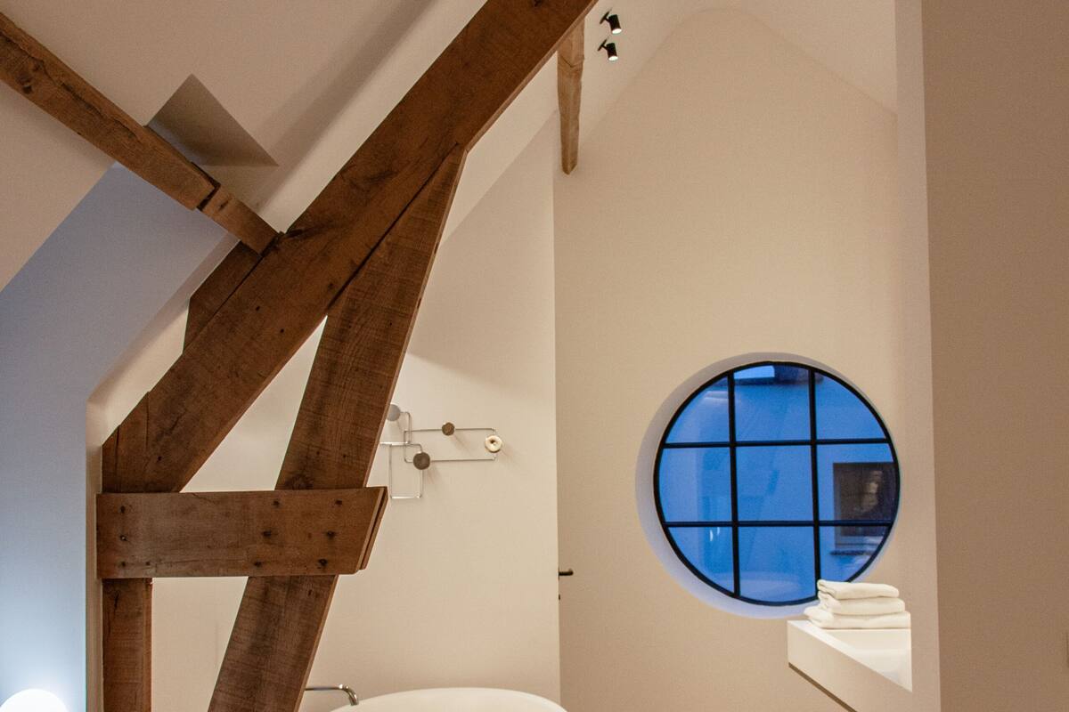 A modern bathroom with a bathtub, a round window, and a wooden ceiling beam.