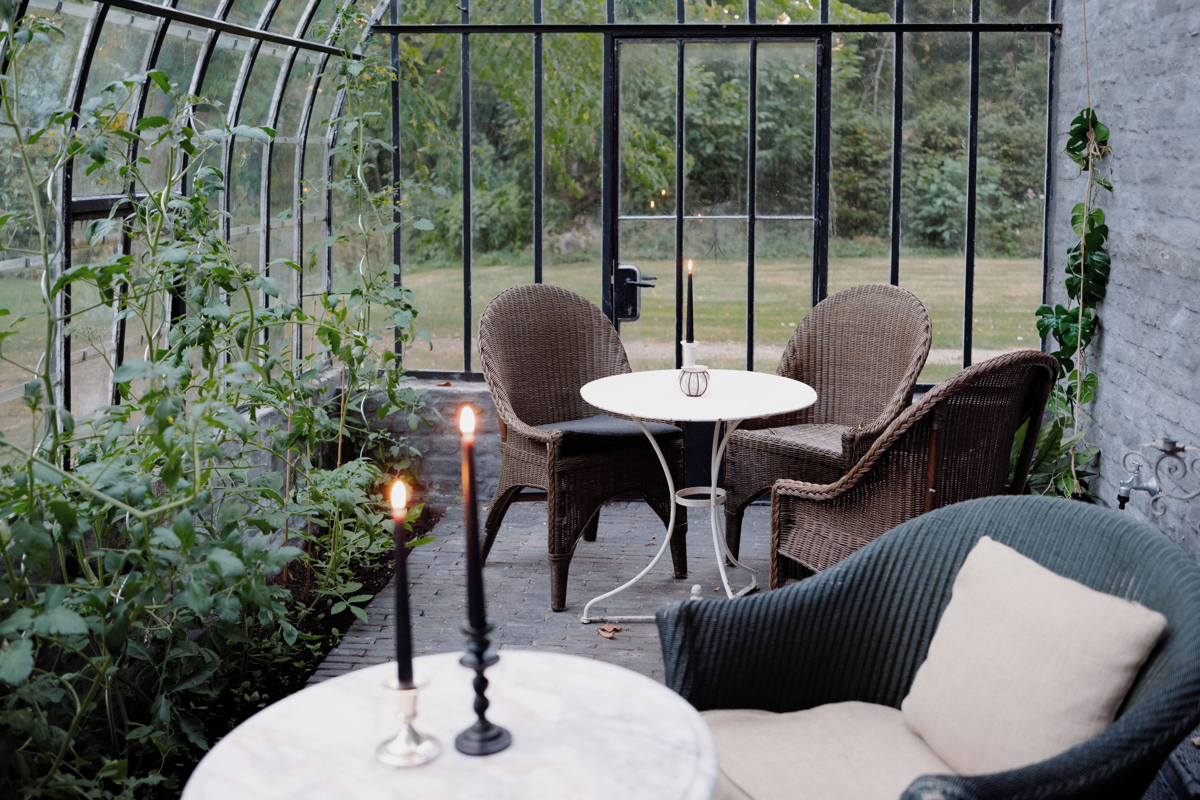A cozy outdoor seating area with wicker furniture and a table, surrounded by greenery and a glass enclosure.