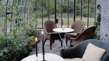 A cozy outdoor seating area with wicker furniture and a table, surrounded by greenery and a glass enclosure.