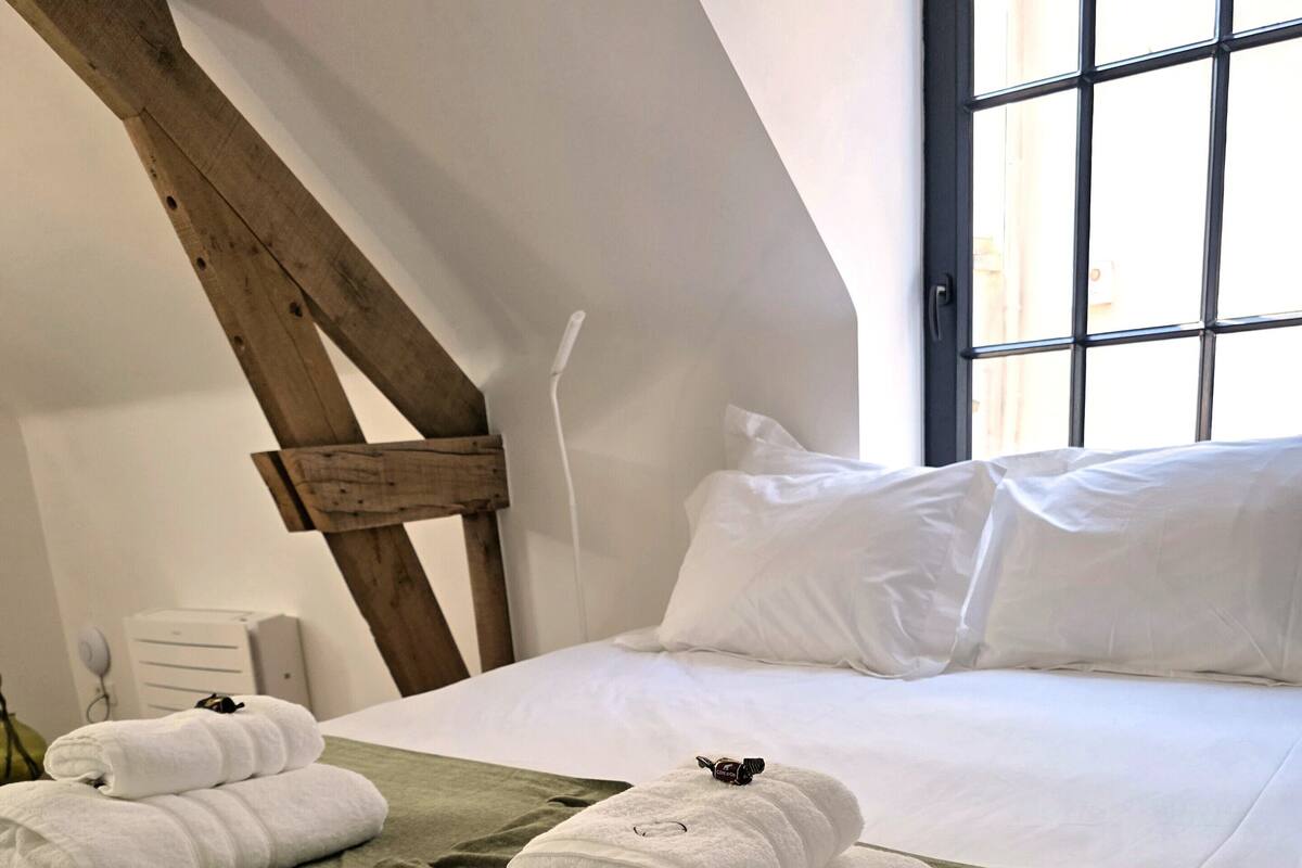 A neatly made bed with white linens and a green bedspread, a wooden beam structure, and a window with a view.