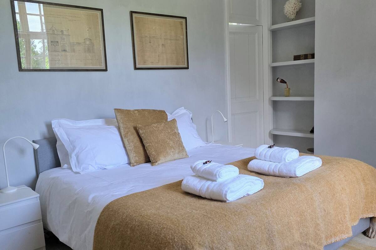 A neatly made bed with white linens and a brown throw blanket, a white bedside table with a lamp, and a shelf with decorative items.