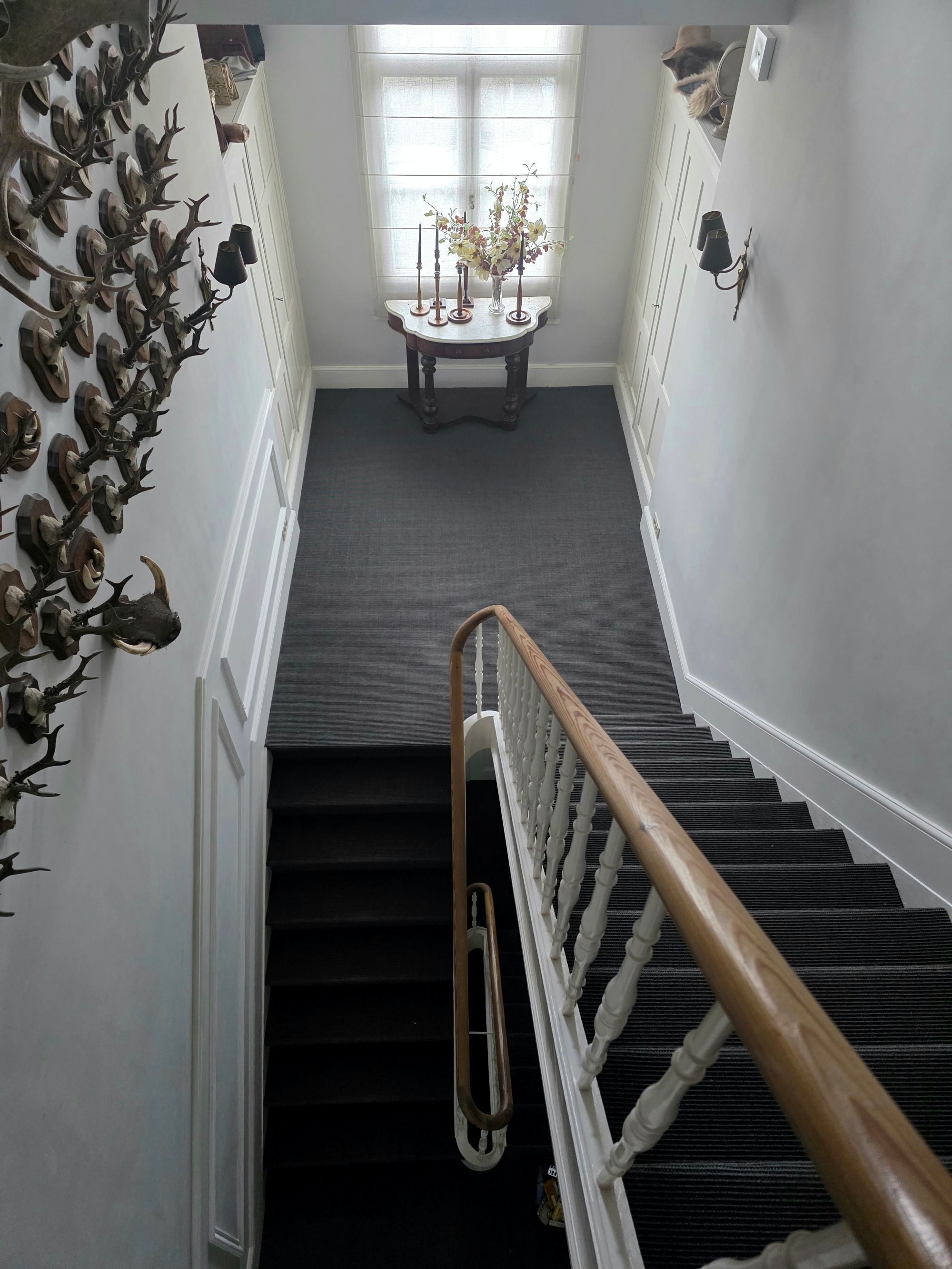 A staircase with a wooden handrail, a wall with mounted antlers, and a table with a vase of flowers in the background.