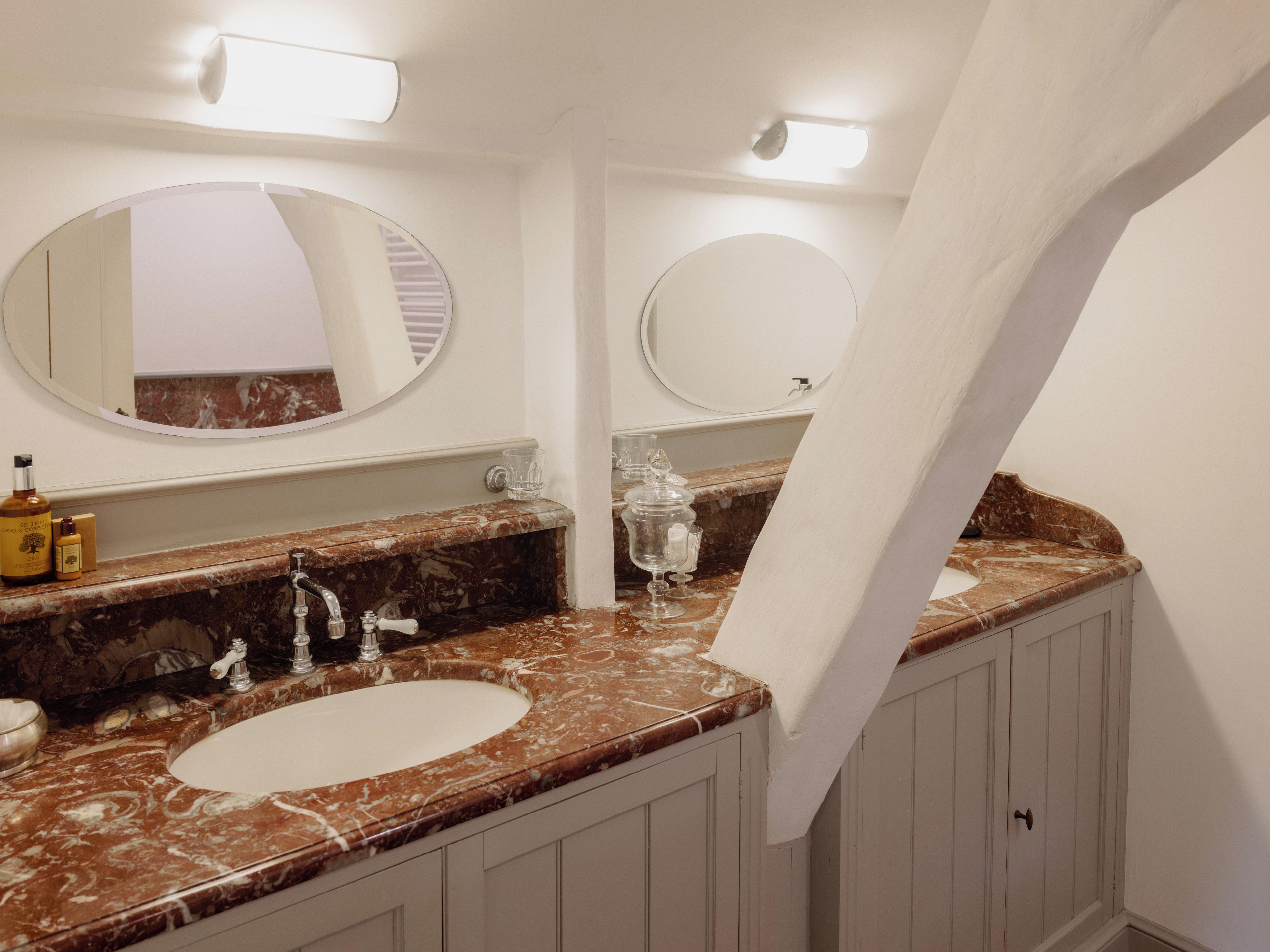 Un baño con dos lavamanos, encimeras de mármol y dos espejos redondos.