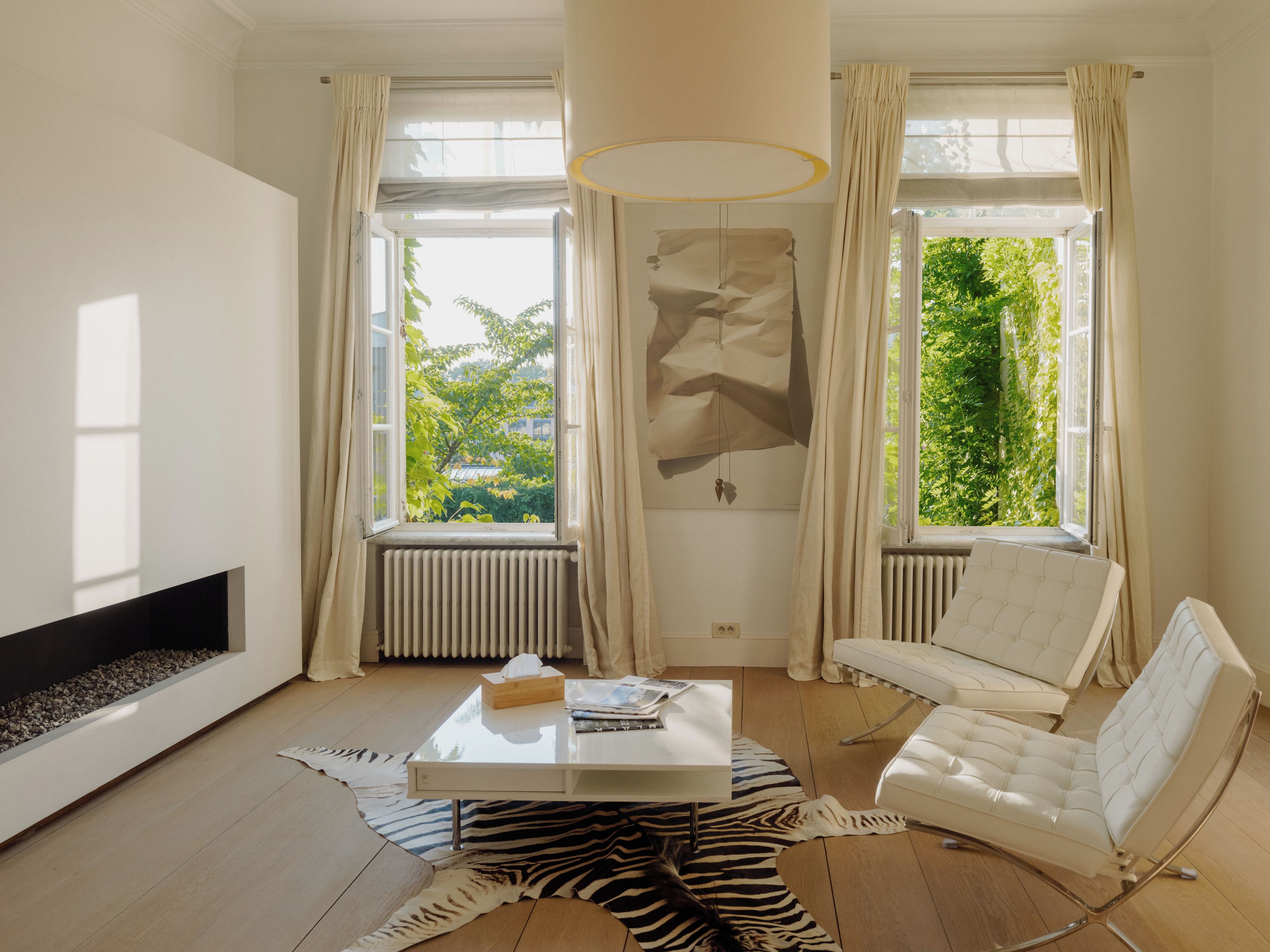 Una sala de estar moderna con chimenea, un sofá blanco, una mesa de centro y ventanas amplias con cortinas.