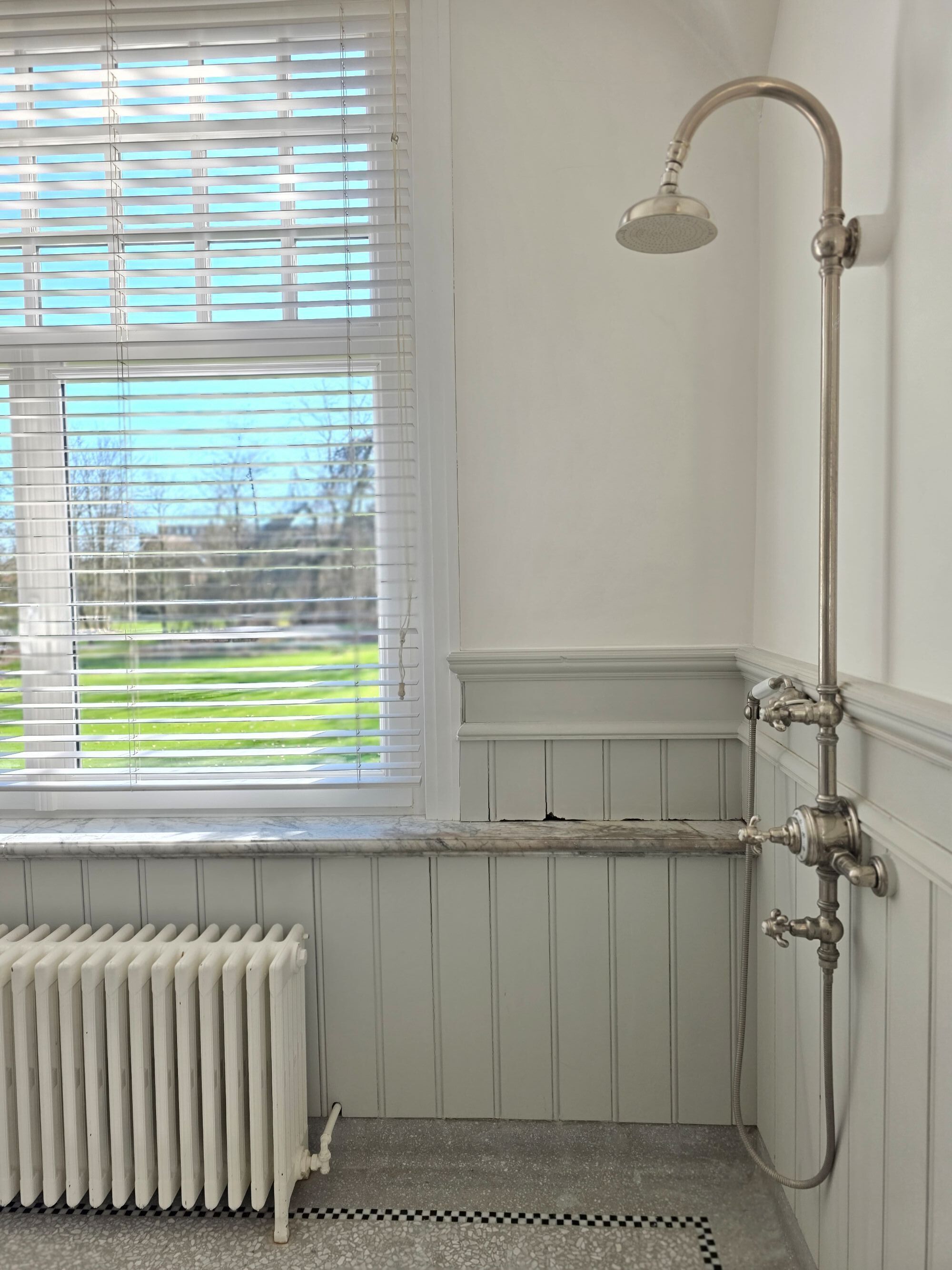 Une salle de bain avec une douche, un radiateur et une fenêtre équipée de stores.