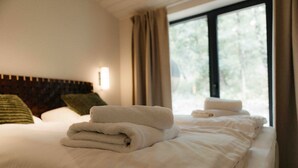 A hotel room with a neatly made bed, a headboard, a window with curtains, and a wall-mounted light.