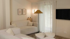 A modern living room with a sofa, two ottomans, a floor lamp, and a flat-screen TV mounted on the wall.