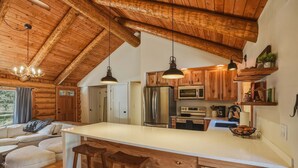 Interior de una cabaña de troncos con cocina, techo de madera y zona de comedor con taburetes.
