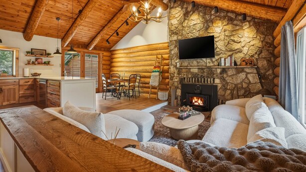 Interior de una cabaña de troncos con chimenea de piedra, mobiliario de madera y televisor montado en la pared.