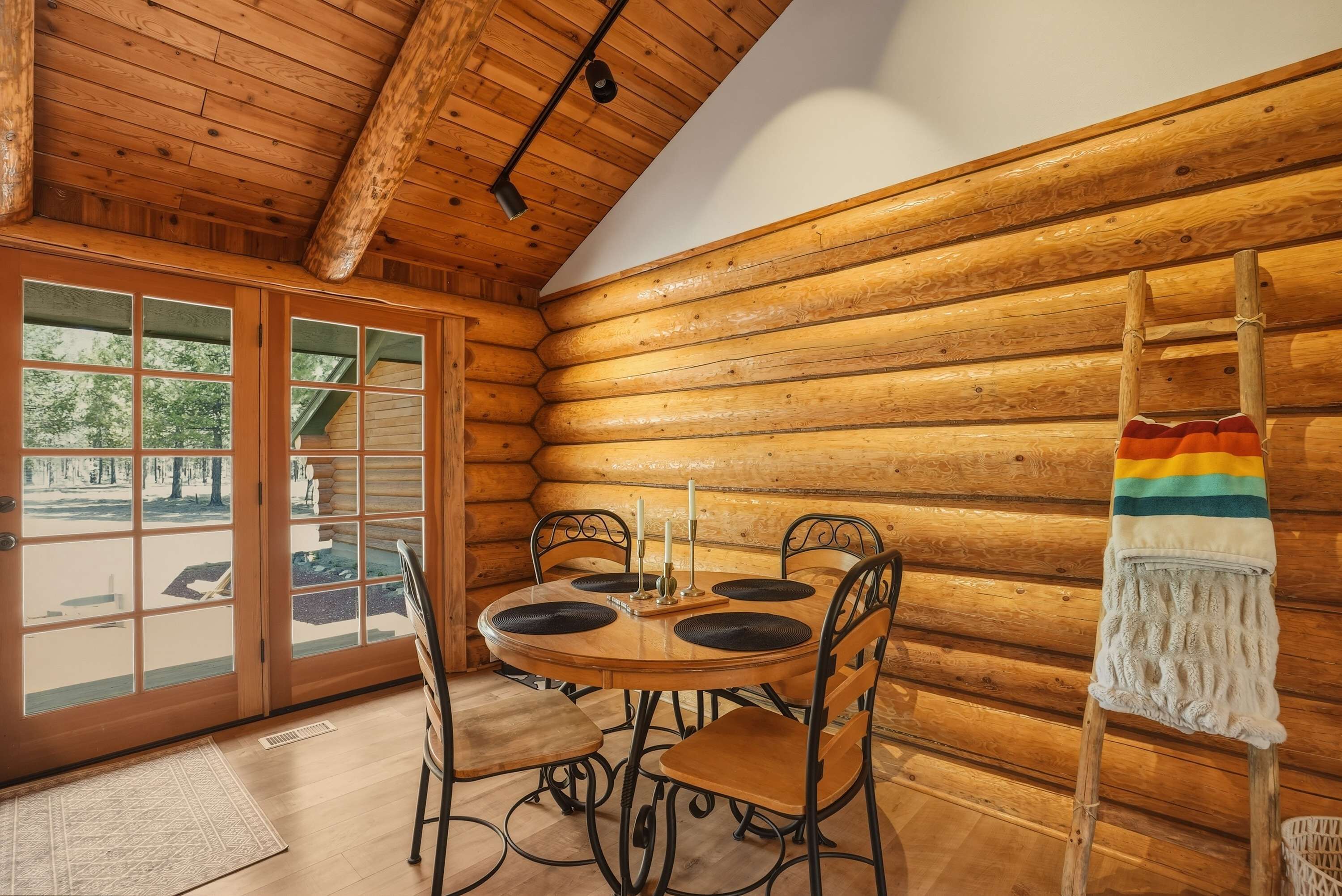 A log cabin interior with a wooden dining table, chairs, a ladder, and a view through a glass door.