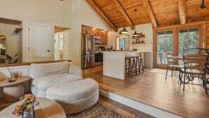 Amplia sala de estar con techo alto, vigas de madera, una cocina con electrodomésticos de acero inoxidable y un comedor con sillas de metal.