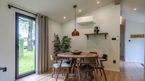 A dining area with a round wooden table, chairs, a hanging pendant light, and a view through a glass door to a garden.