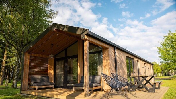 A modern wooden cabin with a covered porch, two sofas, a dining table, and a hammock.