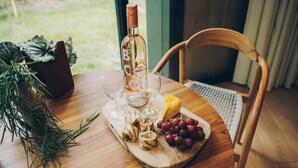 A wooden table with a bottle of Rose Holum wine, two glasses, a cheese wheel, a wooden board with grapes and a wrapped item, and a chair.