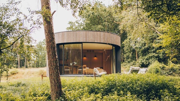 A modern, circular wooden cabin with a glass facade, surrounded by lush greenery.