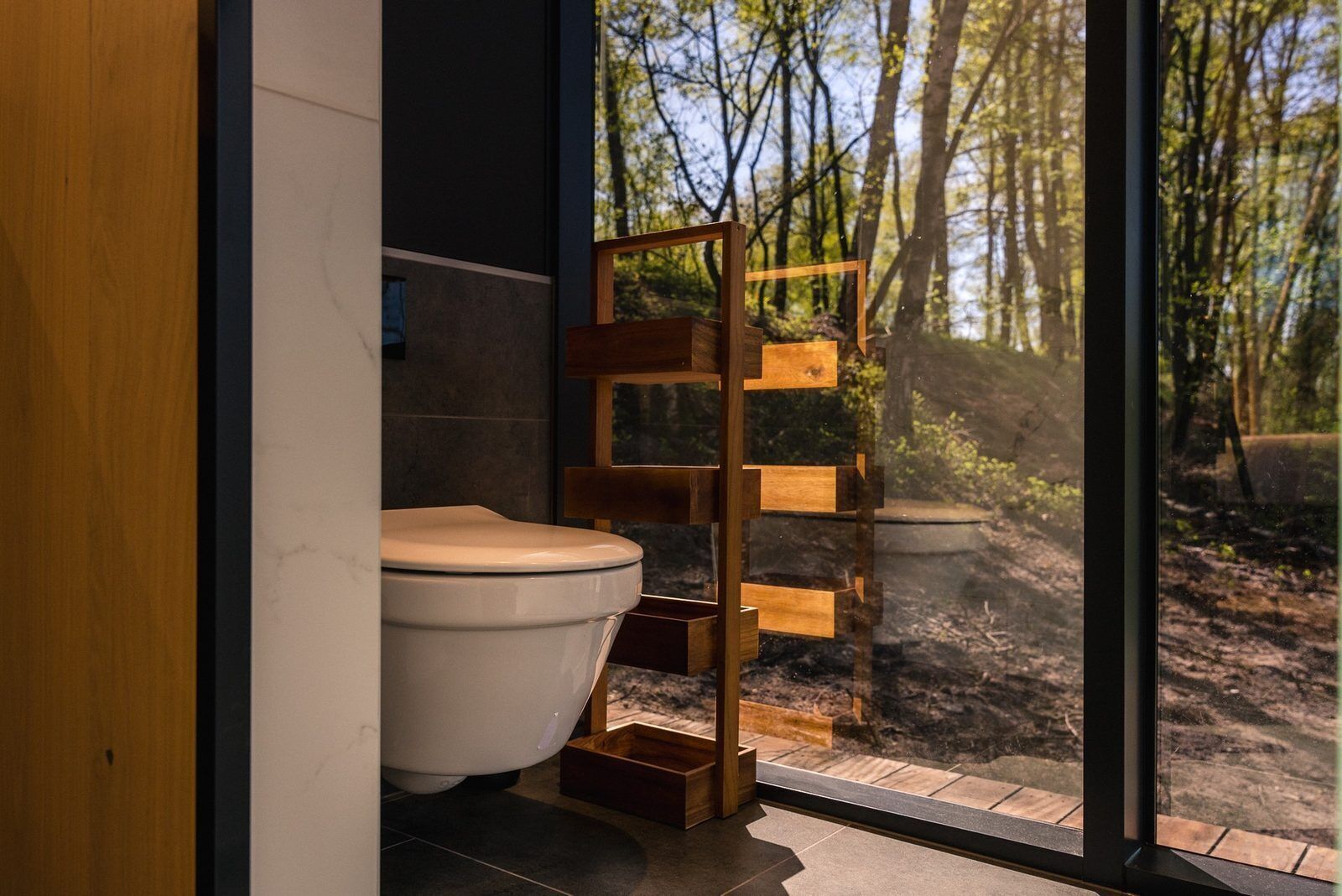 A modern bathroom with a toilet, a wooden shelf, and a large window offering a view of a forest.