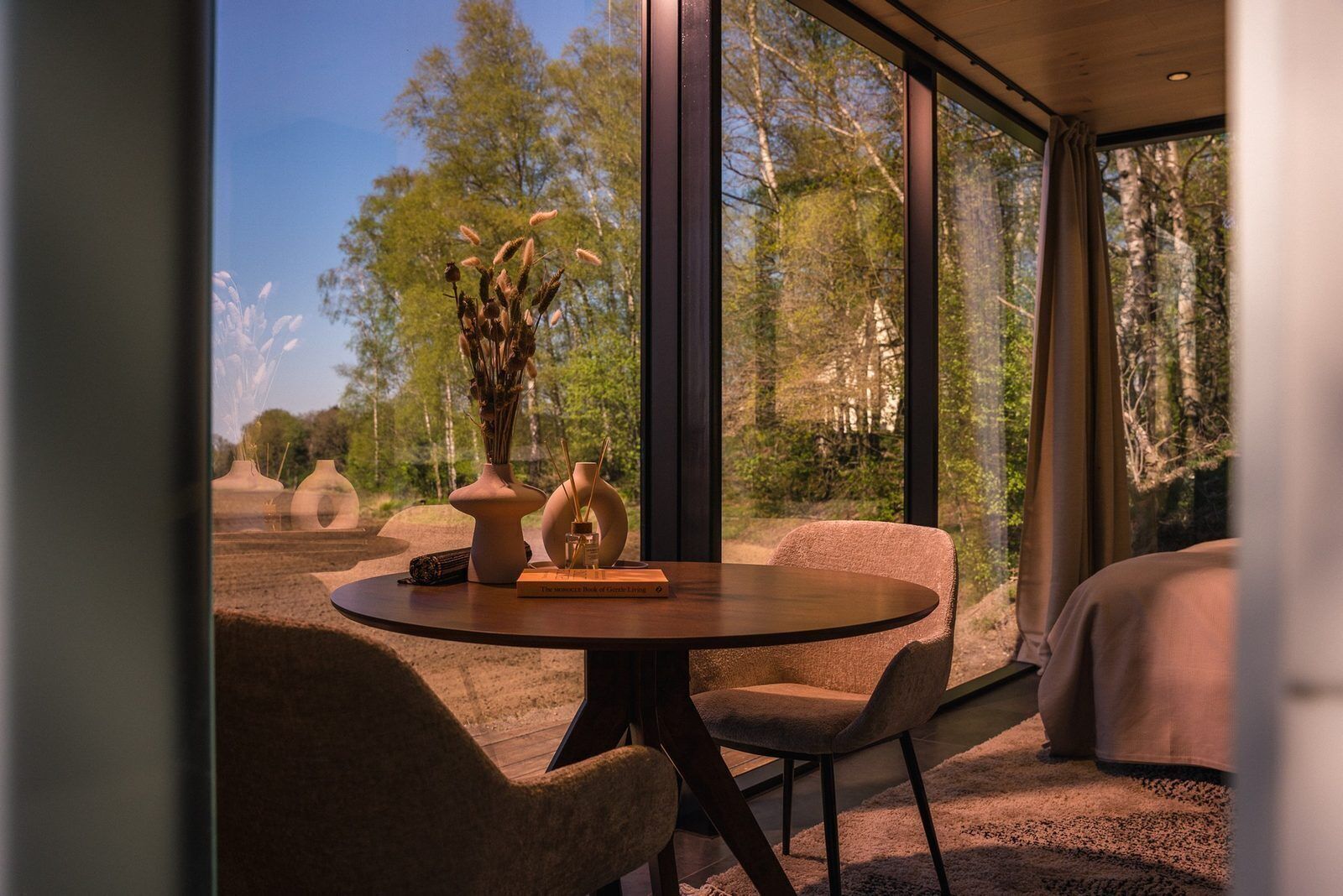 A modern room with a round table, two chairs, a vase with dried flowers, and a book titled 'The Art of Living.'