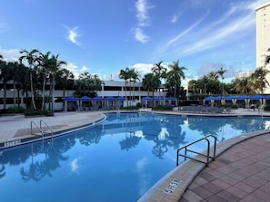 Ein Swimmingpool mit Liegestühlen und Sonnenschirmen, umgeben von Palmen und einem Gebäude mit flachem Dach.