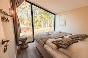Un dormitorio con un ventanal grande, una cama con almohadas, una mesita de noche y una lámpara fijada a la pared.