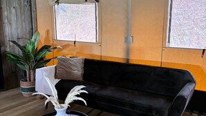 A room with a black sofa, a round coffee table, a potted plant, and two windows with blinds. - Wilsumer Berge Resort - Glamfield Lodge 4p (Wilsum)