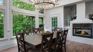 A dining area with a wooden table and chairs, large windows, and a fireplace. - Modern Marble - Big Canoe, GA (Jasper)