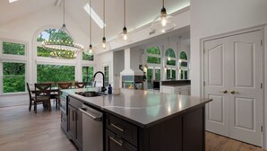 A spacious kitchen with a large island, pendant lighting, and a view of greenery through large windows. - Modern Marble - Big Canoe, GA (Jasper)
