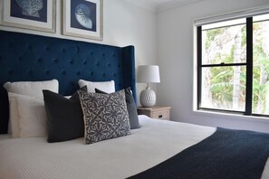 A bedroom with a tufted headboard, a bed with white bedding, patterned pillows, a nightstand with a lamp, and a window with a view of greenery. - Blue Belle - Walk to Beach, Golf & Cafe`s, slps 7 (Shelley Beach)