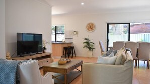 A living room with a TV, sofa, coffee table, and dining area with chairs. - Blue Belle - Walk to Beach, Golf & Cafe`s, slps 7 (Shelley Beach)